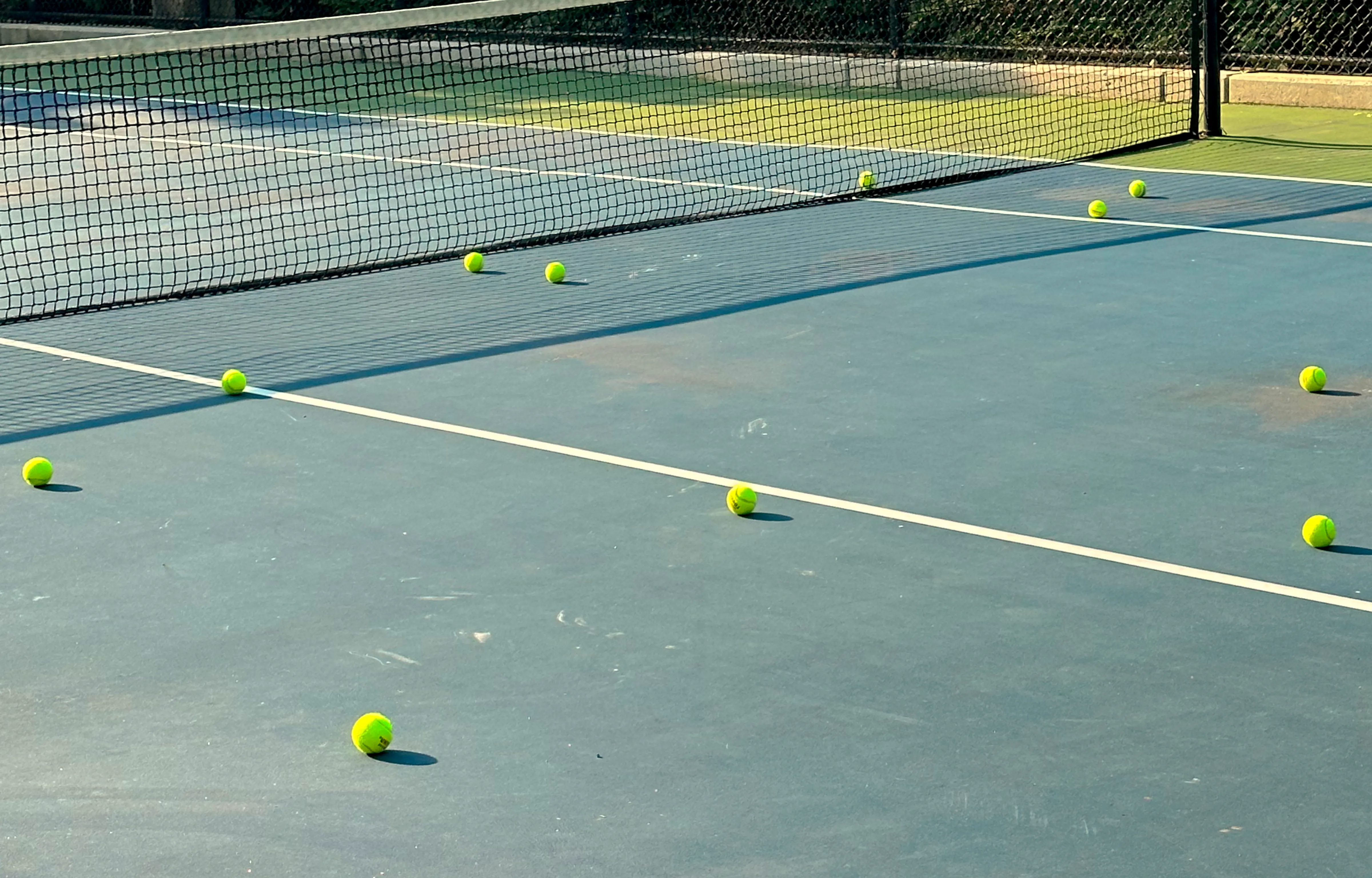 A tennis court with multiple bright yellow tennis balls scattered near the net, indicating a practice session. The court has a blue surface with white boundary lines, and a black net stretches across the middle.